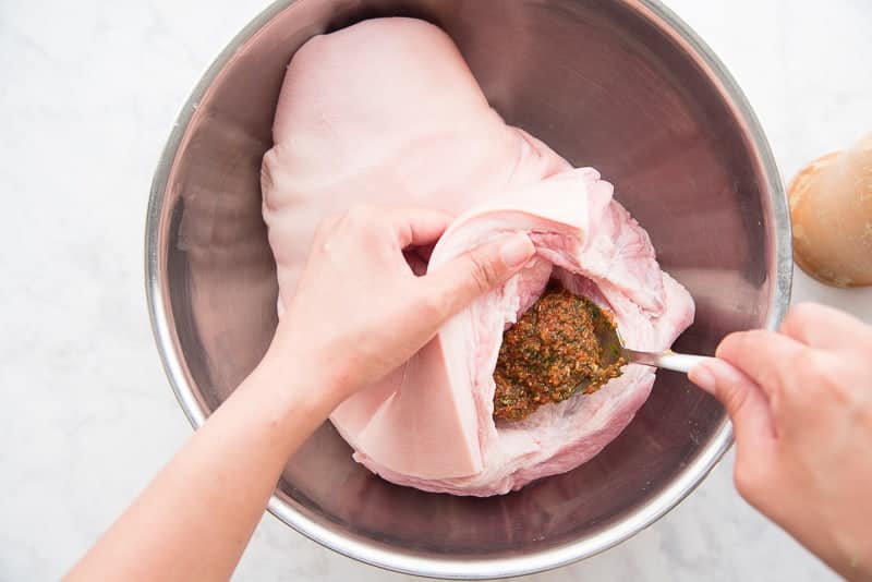 A hand lifts the flap of skin from the pork shoulder while the other hand fills the cavity with the seasoning base from a spoon