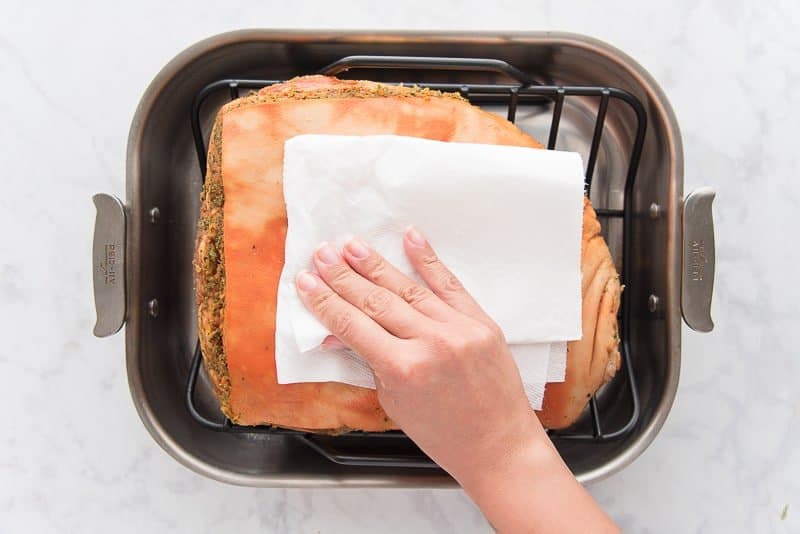 A hand pats the pernil skin with a white paper towel. The pernil is in a silver roasting pan.