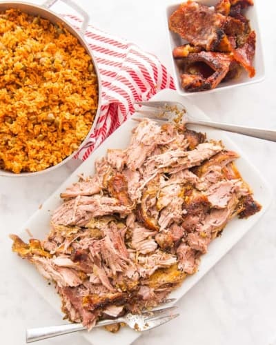 A Puerto Rican dinner of arroz con gandules in a silver pot and shredded pernil on a white platter. A white bowl of chicharron is next to a white and orange striped towel.