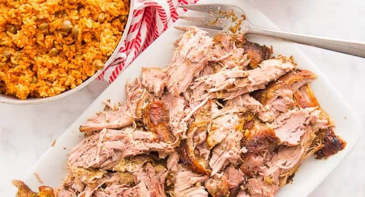A Puerto Rican dinner of arroz con gandules in a silver pot and shredded pernil on a white platter. A white bowl of chicharron is next to a white and orange striped towel.