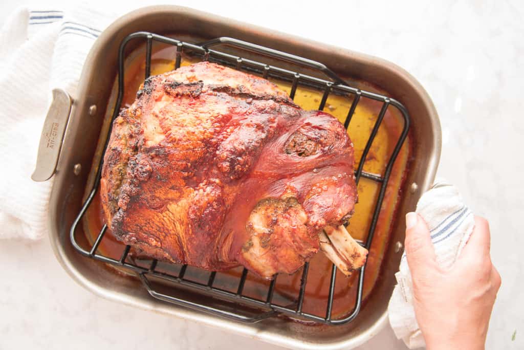 The finished pernil is in a roasting pan. A hand grips the pan with a white and blue towel preview