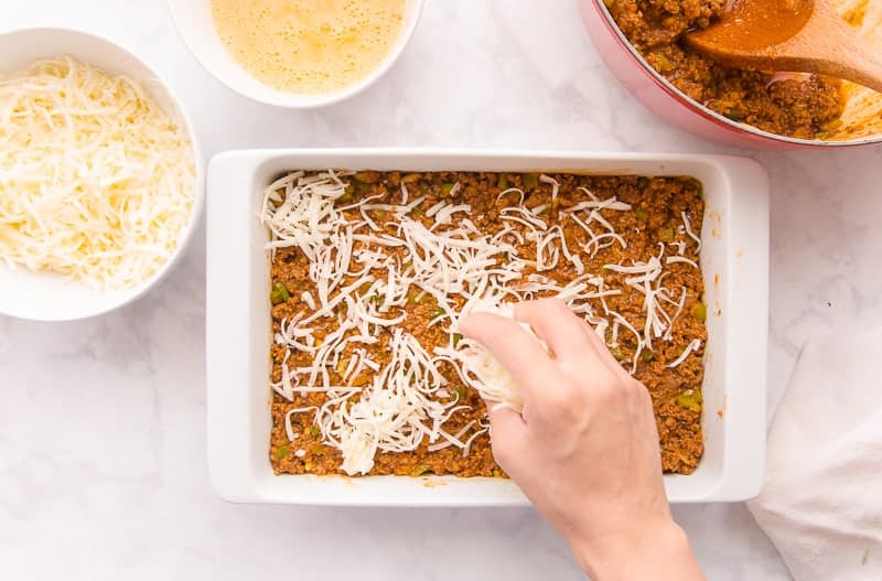Hand sprinkling a layer of shredded mozzarella on the picadillo in a white, rectangular baking dish