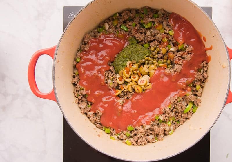 Red tomato sauce, sliced olives, sofrito, and capers are added to a red pot of cooked ground meat.