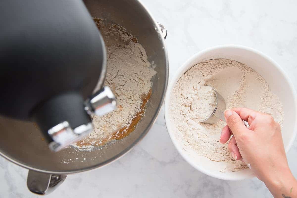 A hand add the flour in a white bowl is added to a silver bowl of wet ingredients. A black mixer.