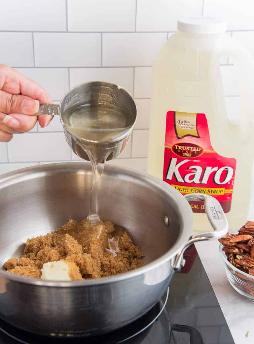 Karo syrup is added to a silver pot filled with brown sugar, nuts, and butter. A gallon jug of Karo syrup is in background.