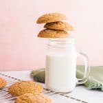 Three brown, soft pumpkin cookies teeter on a glass jug full of milk. Pink background white marble surface. A silver rack has more cookies scattered on it. A green napkin is furled next to the glass of milk.