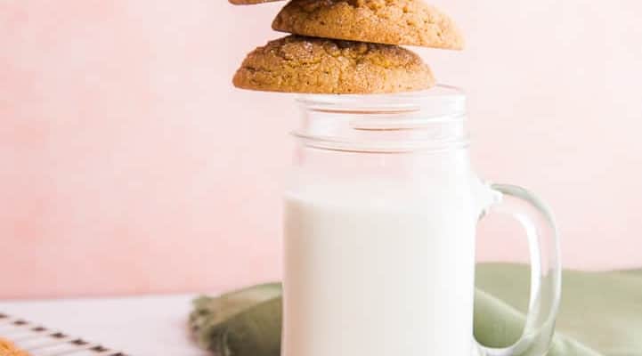 Three brown, soft pumpkin cookies teeter on a glass jug full of milk. Pink background white marble surface. A silver rack has more cookies scattered on it. A green napkin is furled next to the glass of milk.
