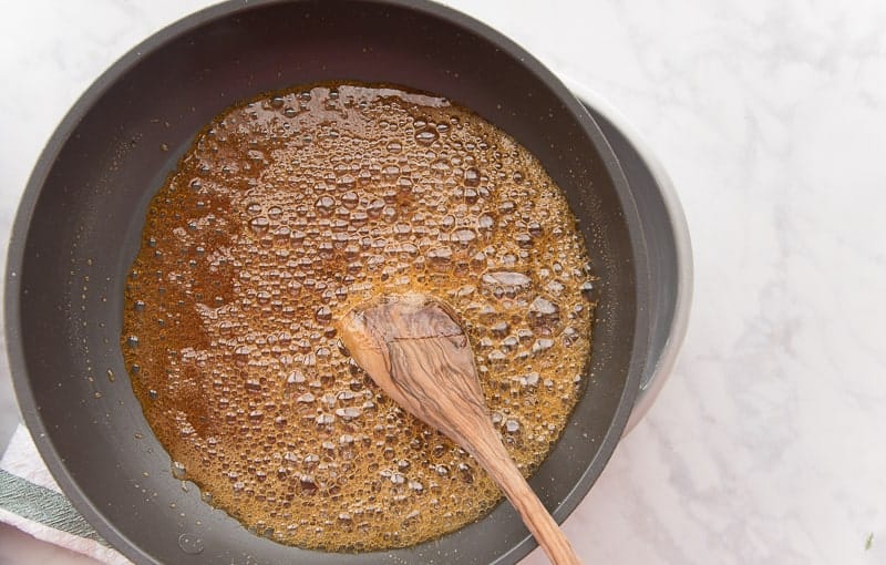Dark brown caramel in a dark-colored pan bubbles while being stirred with a brown, wooden spoon