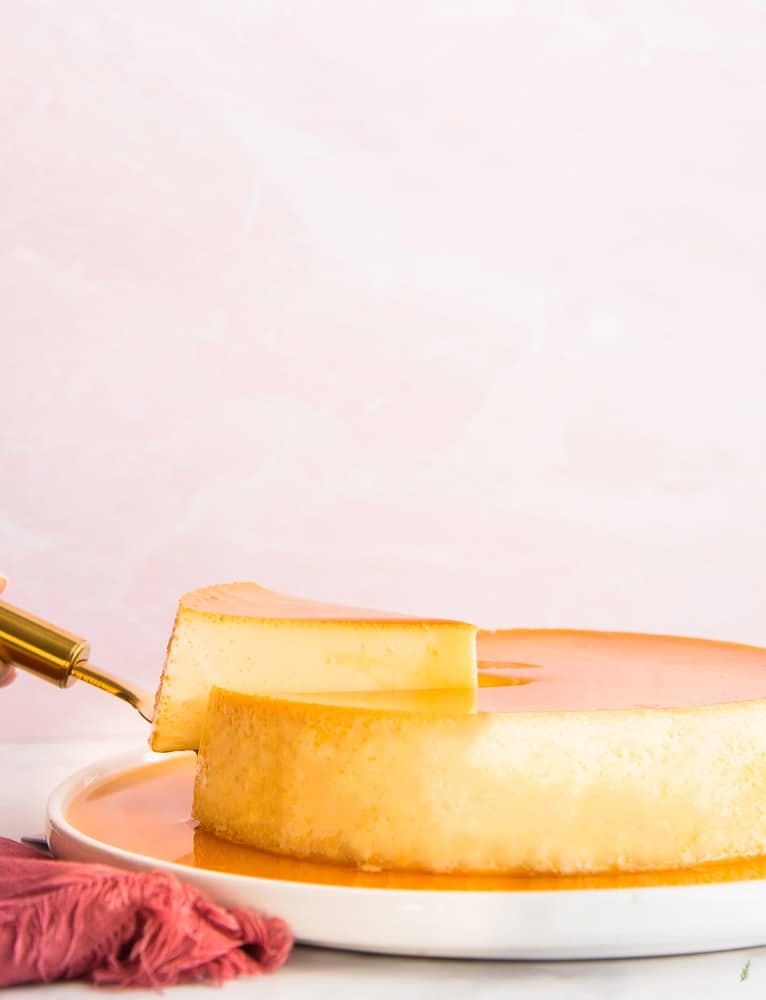 Portrait of a slice of yellow and orange flan being lifted from a whole flan on a round, white plate in front of a pink background. A dark pink napkin furled on left.