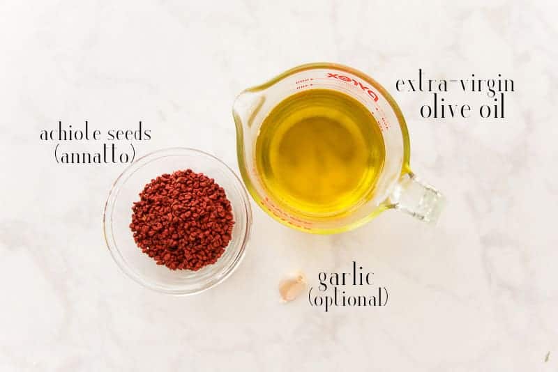 Ingredients to make achiote oil: extra-virgin olive oil, achiote seeds, and garlic are displayed on a white surface
