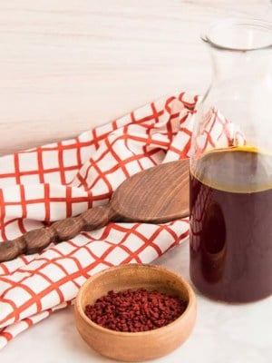 Horizontal image wooden bowl with orange achiote seeds next to a glass bottle half-filled with achiote oil in front of an orange and white windowpane kitchen towel with a wooden spoon on top