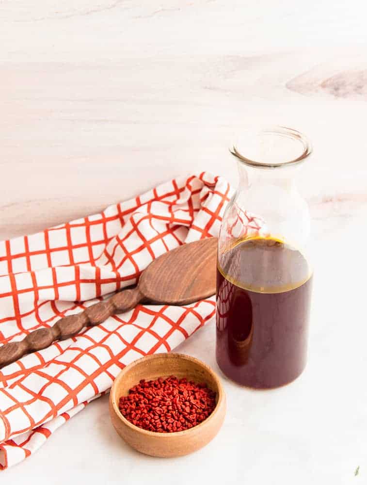Lead image of Achiote Oil: wooden bowl with orange achiote seeds next to a glass bottle half-filled with achiote oil in front of an orange and white windowpane kitchen towel with a wooden spoon on top