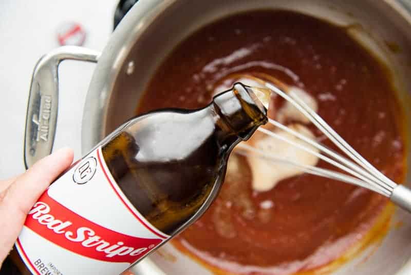 Red Stripe beer is poured from the bottle into the sauce in a silver pot