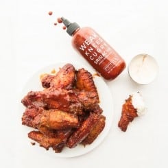 Preview image of a plate of Curry Sriracha Air Fryer Wings next to a spilled bottle of sauce and a single wing dipped in dressing from a silver cup above it