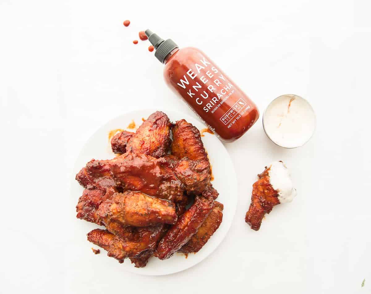 Preview image of a plate of Curry Sriracha Air Fryer Wings next to a spilled bottle of sauce and a single wing dipped in dressing from a silver cup above it