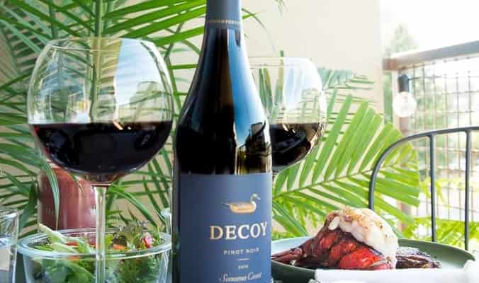 A bottle of Decoy wine sits on a black table in between two glasses of wine and two green plates with a steak and lobster tail on each.