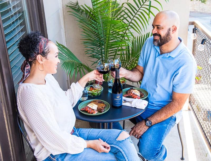 A couple sits at a cafe table on a balcony and toasts over surf and turf dinner