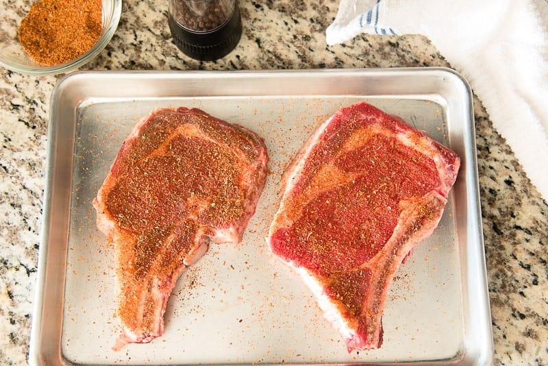 Two ribeye steaks on a silver sheet pan are seasoned. 