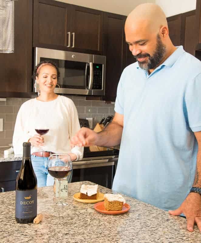 A man and woman in a kitchen. Woman is drinking red wine and talking. Man is sprinkling cinnamon over slices of cake.