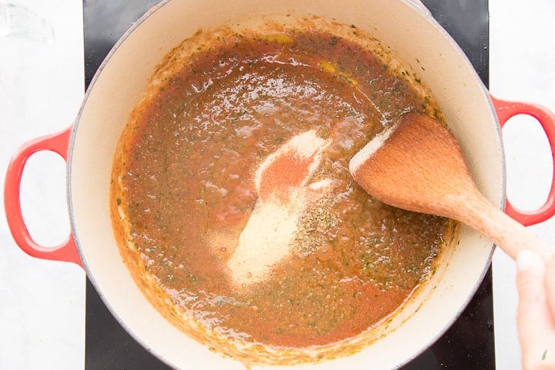 A wooden spoon is being used to stir spices into tomato sauce in a pot