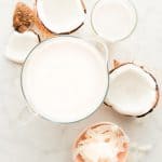 Overhead image of a pitcher of Coconut Milk, a coconut cracked open, a pink bowl with coconut silvers and a glass of coconut milk
