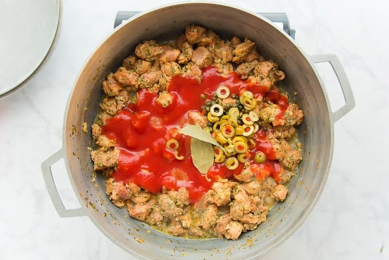 Green olives, capers, bay leaves, and tomato sauce is added to the diced pork for pasteles in a silver pot.