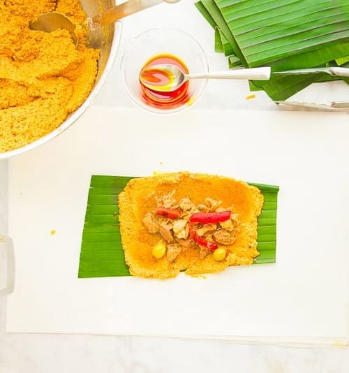 orange masa is on a green banana leaf and covered in pork filling, red pimentos, and two green olives