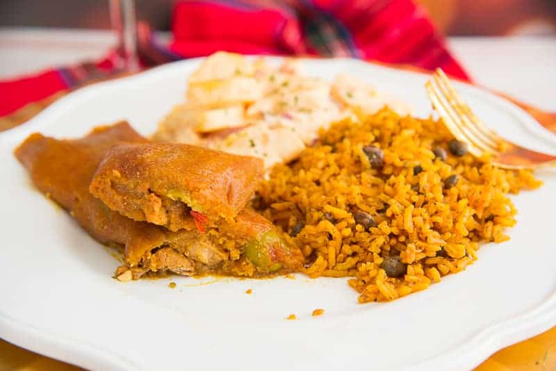 A brown pastel with pork filling exposed on a white plate with yellow rice and potato salad
