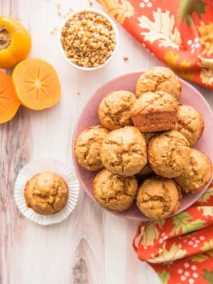 Long image pink plate with muffins. One muffin in a white wrapper on a wooden surface. A white bowl of chopped nuts.An orange persimmon cut in half leans against a whole fruit.