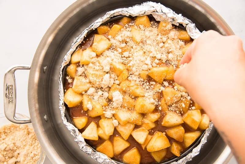 A hand sprinkles the streusel on the apple pie topping on the apple cheesecake