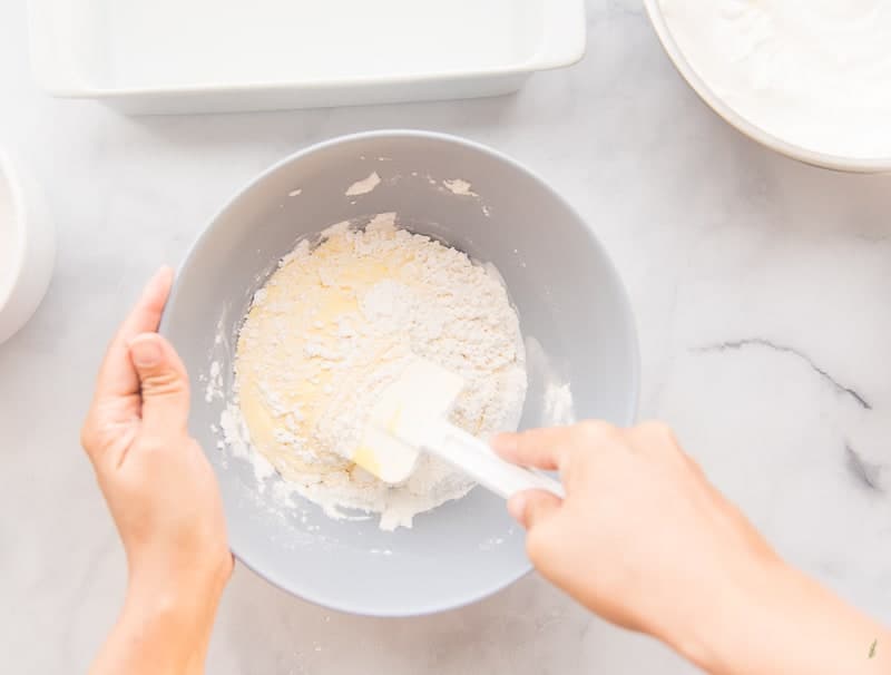 A hand holds a blue mixing bowl while the other hand folds flour into a yellow egg batter.