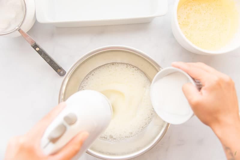 A hand gradually adds sugar to a silver mixing bowl of whipping egg whites