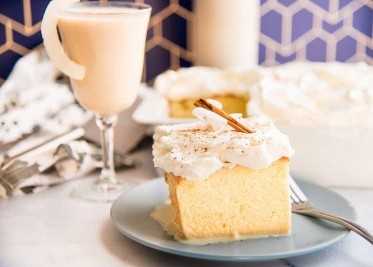 A slice of cake topped with whipped cream and a cinnamon stick on a blue plate, accompanied by a fork. In the background, a glass filled with a creamy beverage and a cake in a dish are visible. The setting features a geometric tiled wall.