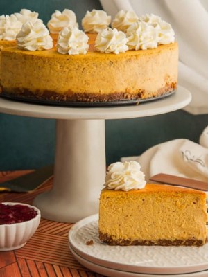 A slice of pumpkin cheesecake on a stack of white plates in front of a gray cake stand with a cheesecake on it