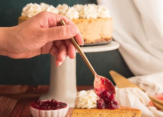 A hand spoons cranberry sauce on a slice of pumpkin cheesecake