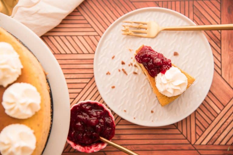 A slice of cheesecake topped with whipped cream and red berry sauce on a decorative plate. A fork rests beside it. A small bowl of berry sauce and part of a larger cheesecake are visible on a wooden textured surface.