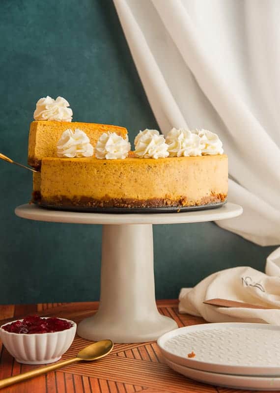 Slice of pumpkin cheesecake is being lifted from the Cheesecake on a gray cake stand