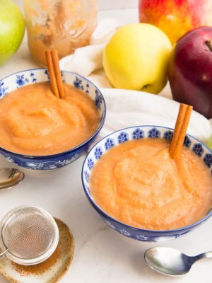 Two blue and white floral bowls of Slow Cooker Cinnamon Applesauce surrounded by apples of different colors and a sifter of cinnamon