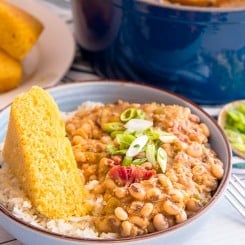 A closeup image of a blue bowl filled with white rice and topped with black eyed peas garnished with sliced green onions. A wedge of yellow cornbread sits on the left