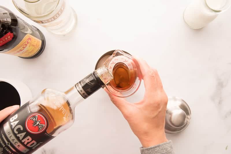 A hand pouring dark rum from its bottle into a jigger over a silver cocktail shaker