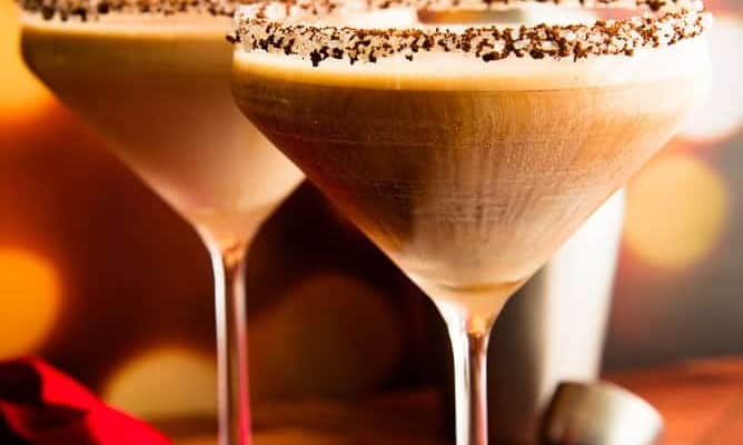 Lead image of Two martini glasses rimmed with ground espresso and sugar crystals and filled Café con Leche Martini on a black slate surface next to a pile of chocolate candies.