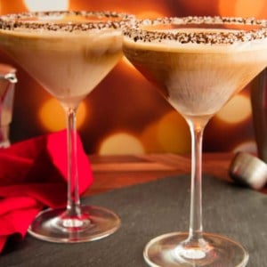 Preview image of Two martini glasses rimmed with ground espresso and sugar crystals and filled Café con Leche Martini on a black slate surface next to a pile of chocolate candies.