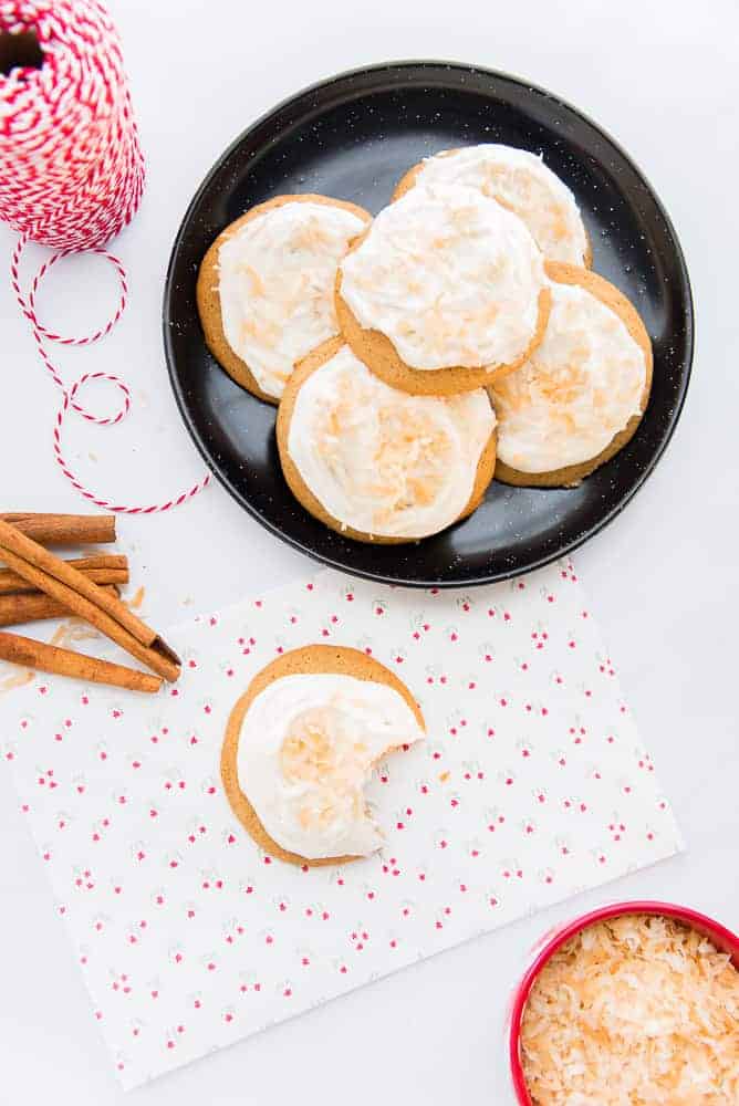 Lead image of coquito cookies. Coquito cookies on a black plate garnished with toasted coconut, a cookie with a bite removed on a white piece of paper with floral print. Bottom right of image: a red bowl filled with toasted coconut. Top-left image: red and white striped spool of string.