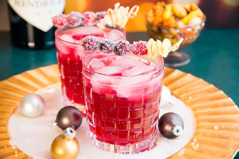 To Ginger Rickey cocktails garnished with sugared cranberries and furled ginger on a gold charger topped with metallic ornaments in sugar crystals