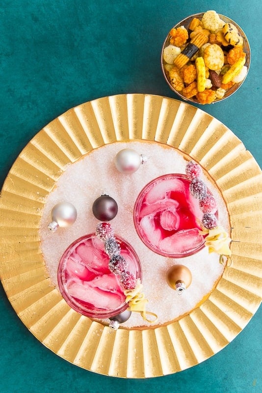 Portrait image of a gold charger topped with white sugar crystals pearl ornaments. Two cocktail glasses filled with Ginger Rickey cocktails garnished with sugared cranberries and furled ginger. Top right of image a bowl of cocktail snacks on a green background.