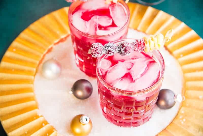 Three-quarter image of two Ginger Rickey cocktails in old-fashioned glasses garnish with sugared cranberries and furled ginger on a gold charger top with white sugar crystals and ornaments