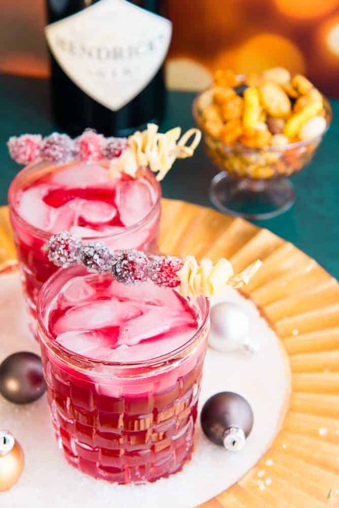 Portrait image of two old-fashioned glasses filled with ginger rickey cocktail garnished with sugared cranberries and furled ginger. Top right background: a bowl of cocktail snacks in a bottle of gin
