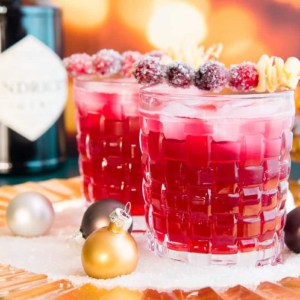 Horizontal image of two old fashioned glasses filled with ginger Rickey cocktail garnished with sugared cranberries and furled ginger.
