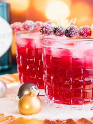 Horizontal image of two old fashioned glasses filled with ginger Rickey cocktail garnished with sugared cranberries and furled ginger.