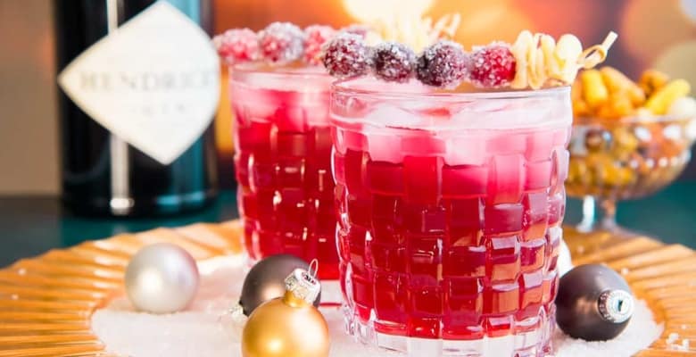 Horizontal image of two old fashioned glasses filled with ginger Rickey cocktail garnished with sugared cranberries and furled ginger.